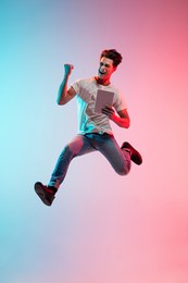 winner with tablet. young caucasian man's jumping on gradient blue-pink studio background in neon light. concept of youth, human emotions, facial expression, sales, ad. full length, copyspace.