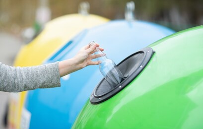 waste sorting and recycling concept. unrecognizable female throwing empty glass bottle into green recycle bin garbage container outdoors, caring about environment, cropped image with selective focus