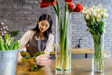 floristics, business, decoration concept. asian female sales assistant working as florist standing behind counter wearing yellow sweater and grey apron with brick grey wall on background.