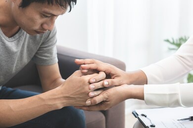 young asian man, male suffers from a mental who needs to therapy with a psychologist while sitting on couch to consult, psychiatrist has encouragement the patient by touching to make his feel relaxed.