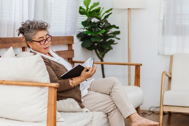 happy healthy retired smiling senior woman or pensioner reading book resting or sitting on couch or sofa at home. elderly adult active lifestyle and activity on holiday. health and insurance concept