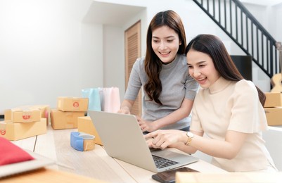 two beautiful asian women are checking orders by laptops via the internet. and pack the paper box with a happy smiling face, being a new normal online business