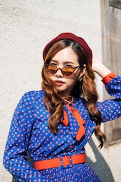 medium portrait shot of wavy brunette woman in blue and red retro dress standing with cool posture at the old house's wall in sunlight.