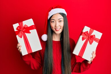 young chinese woman wearing christmas hat and holding gifts smiling with a happy and cool smile on face. showing teeth. 