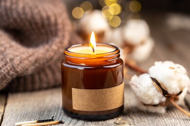burning candle in a glass jar with empty label mock up. cosy and soft winter background, knitted scarf and brunch of cotton flower. christmas holidays at home.