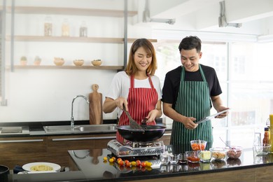 young asian man in green apron watches his girlfriend prepare meal. couple enjoy cooking  in kitchen. home activities. quarantine concept copy space.