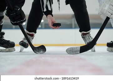 hand of referee holding puck over ice rink with two players with sticks standing around and waiting for moment to shoot it