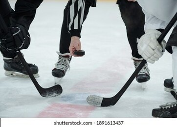 hand of referee holding puck over ice rink with two hockey players with sticks standing around him and waiting for moment to shoot it