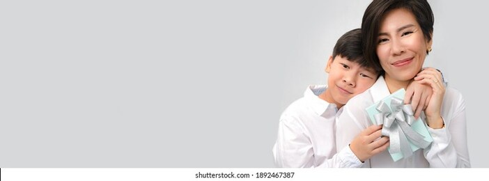 a happy and healthy smart looking asian preteen boy embracing his beautiful mom with present on mother's day or birthday. love expression, parenting teenager, mother and child, young big boy. banner.