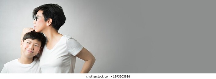 a smart and beautiful asian mom holding her preteen son with love. her son leans his head on her chest smiling happily. love expression, parenting teen, single mom, young, mother love concept banner.