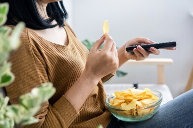 woman hand eating potato chips and holding remote tv watching series sitting on sofa 