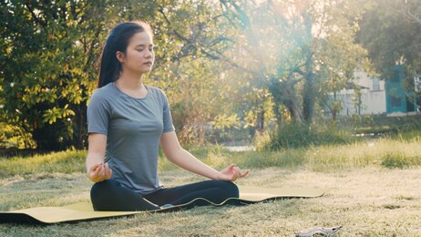 attractive asian woman sitting in meditation pose position yoga pranayama balance in the beautiful the park view during sunset. health and wellness concept.
