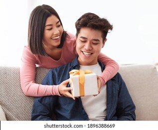asian wife giving wrapped gift to husband surprising him on valentine's day sitting on sofa at home. family holidays celebration, st valentine presents and gifts concept