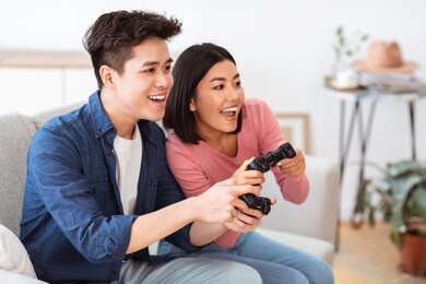 happy asian couple playing video game having fun together on weekend sitting on couch at home. computer gaming concept. millennials friends play videogames indoors. side view