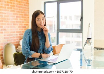 young asian woman looking happy and smiling with hand on chin, wondering or asking a question, comparing options