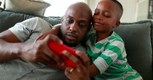 father and child together watching content on smartphone device. african little boy embracing parent