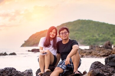 asian couple sitting on the rocks by the beach with the sunset at chonburi province, thailand.