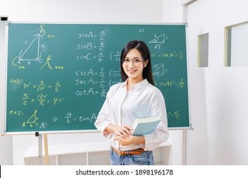 asian senior high school female math teacher hold books and smile at you in front of blackboard at classroom
