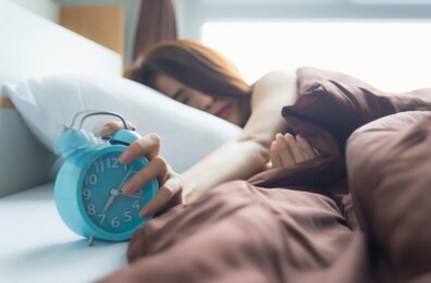 young asian woman sleeping on bed pressing snooze button on blue  alarm clock at seven o'clock morning in bed room at home, lifestyle, good morning, healthy sleep and joyful weekend concept