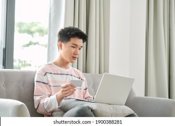 profile view of young mixed ethnicity man sitting on sofa at home with laptop computer using credit card to shop online
