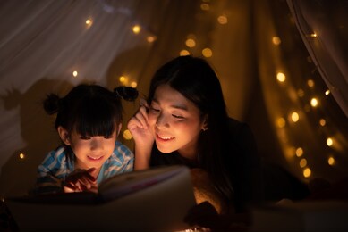 young asian mother read the story in the book for her daughter children on the bed at home, family and motherhood learning concept