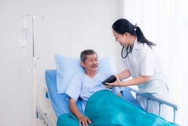 asian female doctor wearing a white gown and stethoscope , aged 40-50 years is describing the illness and treatment for an elderly male patient in a hospital bed with a tablet