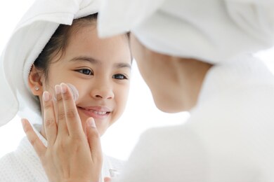 mother adding treatment cream on the cheek to young and cute asian girl with spa dress and head covered with a white towel.