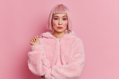 serious pretty asian woman with trendy pink hair dressed in winter coat has bright vivid makeup poses against rosy background. teenage girl dresses for walking during cold weather. fashion concept