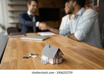 focus on close up keys bunch and cottage house toy model on wooden table. smiling young man broker realtor real estate agent shake hands of happy black couple clients homeowners on blurred background