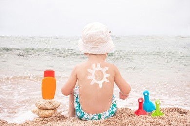 the sun drawing sunscreen (suntan lotion) on baby (boy)  back. caucasian child is sitting with plastic container of sunscreen and toys on beach. close up, outdoor, copyspace (sharm el sheikh, egypt). 