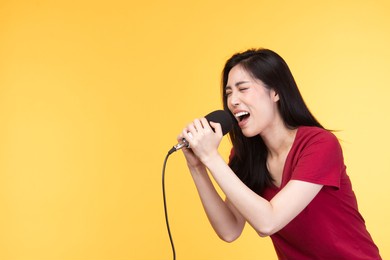 pretty asian woman in casual wear holding microphone and singing on yellow background.