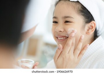 mother adding treatment cream on the cheek to young and cute asian girl with spa dress and head covered with a white towel. the kid's face expresses a happy smile and joy.