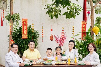 happy big asian family sitting at big dinner table outdoors and celebrating lunar new year, decorations with best wishes inscription in background