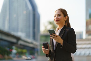 asian executive working woman holding coffee cup and using a mobile phone in the street with office buildings in the background in bangkok, thailand.
