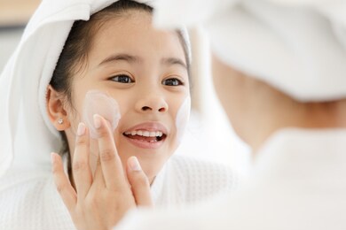 mother adding treatment cream on the cheek to young and cute asian girl with spa dress and head covered with a white towel. the kid's face expresses a happy smile and joy.