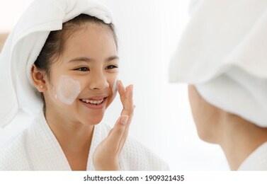 mother adding treatment cream on the cheek to young and cute asian girl with spa dress and head covered with a white towel. the kid's face expresses a happy smile and joy.