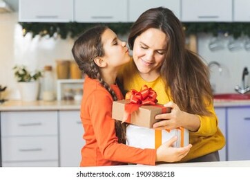 happy family: daughter kisses her mother a gives gift and flowers for the holiday. a cute girl in the kitchen gives her mom a box and hugs for mothers day