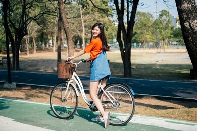 long hair girl in orange t-shirt and jeans jumper stop her bike in the bicycle lane at the park and smile to the camera.