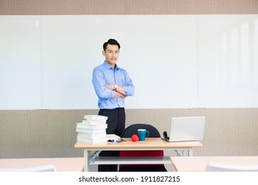 asian young male teacher smiles at you with cross arm standing front of whiteboard in classroom