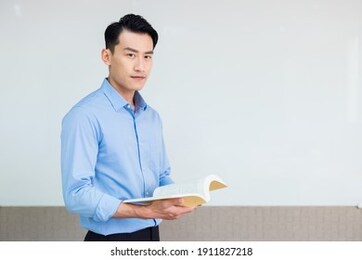 asian young male teacher holds a book and smiles at you standing front of whiteboard in classroom
