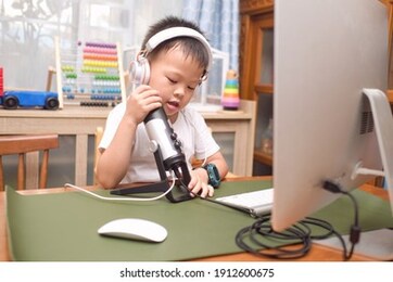 asian 5 years old boy child wearing headphones using microphone with computer prepare to making video call to relatives at home or making vlog for social media channel, distance learning concept