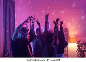 young intercultural friends in smart casualwear raising arms while dancing together at home party in living-room illuminated with pink lighting