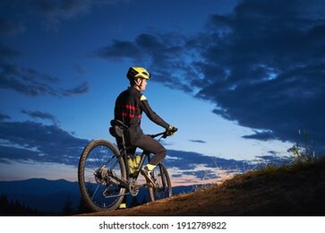 back view of young man in cycling suit sitting on bicycle under blue night sky with clouds. male bicyclist in safety helmet riding bicycle on the road in the evening. concept of active leisure.