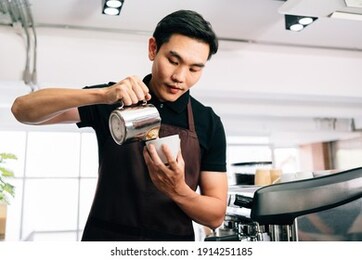 a young asian barista man in an apron dress intentionally poured hot milk into a hot espresso black coffee for making latte art.