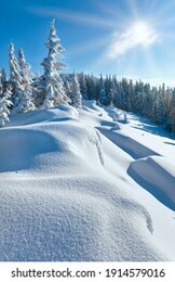 snowdrifts on winter snow covered mountainside, fir trees on hill top and sun shine in blue sky