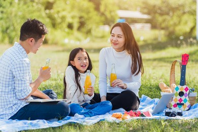 happy asian young family father, mother and child little girl having fun and enjoying outdoor sitting on picnic blanket drinking orange juice from glass bottle, summer resting at a nature garden park