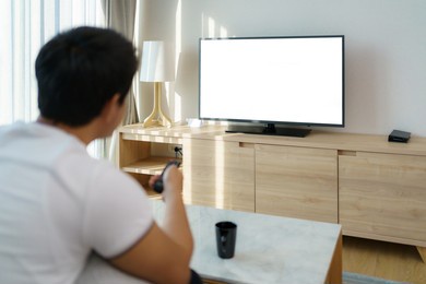 an asian man holds a television remote and is pressing the channel while watching tv on the couch in the living room at home.