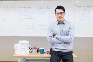 asian young male teacher smiles at you with cross arm in classroom