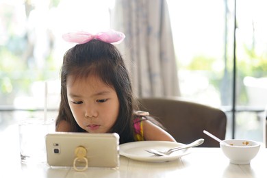 asian child or kid girl enjoy eating breakfast and watch smartphone or mobile phone to e-learning communication or study online and people learn from home or play game and video clip by back to school