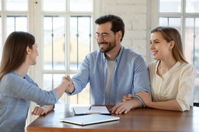 happy young married family couple shaking hands with real estate agent, getting acquainted at meeting, celebrating making agreement or thanking for high quality professional service in office.